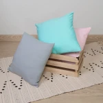 Poszewka jersey 40x40 w kolorze popielatym leży na podłodze obok kolorowych poduszek w odcieniach miętowym i różowym, umieszczonych w drewnianym koszyku. Tło jest jasne, a poduszki mają gładką teksturę. Widać także tkaninę z wyraźnym wzorem.
