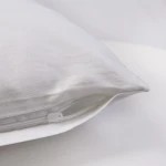 Poszewka satynowa 70x80 w kolorze białym, leżąca na jednolitym tle. Wykonana z gładkiego materiału z zamkiem błyskawicznym, co pozwala na łatwe zakładanie i zdejmowanie. Szczegół widać w dolnym rogu poszewki.