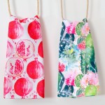 Komplet dwóch ścierków kuchennych Tropical Fruits w rozmiarze 38x60 cm. Po lewej stronie ścierka z czerwonymi owocami granatu, po prawej zielona z motywem tropikalnych roślin, zawieszone na sznurku.
