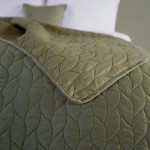 Narzuta Verde mink w kolorze zielonym, o wymiarach 170x210 cm, leży na białej pościeli. Zbliżenie ukazuje fakturę tkaniny z delikatnym wzorem liści. Widoczna poduszka w tym samym kolorze uzupełnia aranżację. Rant narzuty ma miękkie wykończenie.