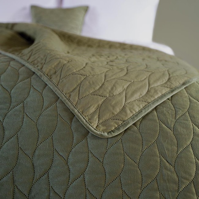 Narzuta Verde mink w kolorze zielonym, o wymiarach 170x210 cm, leży na białej pościeli. Zbliżenie ukazuje fakturę tkaniny z delikatnym wzorem liści. Widoczna poduszka w tym samym kolorze uzupełnia aranżację. Rant narzuty ma miękkie wykończenie.