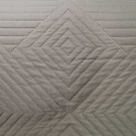 Narzuta dwustronna Lamia w kolorze szaro-ecru, wykonana z miękkiego materiału. Widoczny detal w postaci wzoru w diagonalne romby oraz prostych linii. Zdjęcie zbliżeniowe, ukazujące teksturę i wykończenie. Brak widocznych napisów lub logo.