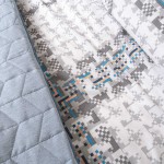 Pościel bawełniana 220x200 Cottonlove w kolorze szarym z wzorem w kratkę. Materiał o gładkiej fakturze, z pikowanym, szarym akcentem po lewej stronie. Produkt rozłożony na płaskiej powierzchni, ukazujący szczegóły wzoru i kolorowe elementy dekoracyjne.