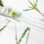 Zbliżenie na zamek błyskawiczny pościeli bawełnianej w kolorze białym z kwiatowym wzorem w odcieniach zieleni i niebieskiego. Produkt leży na płaskiej powierzchni, ukazując detale materiału oraz sposób zamknięcia poszewki.