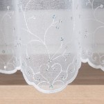 Firana haftowana w delikatne kwiaty, biała, wysokość 160 cm. Zbliżenie na dolną krawędź, ukazujące haftowane wzory i małe kryształki. Tło jasne, jednolite, co podkreśla detal oraz subtelność materiału. Firana wisząca, zwiewna, idealna do aranżacji okien.