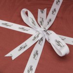 Zbliżenie na prezent zapakowany w ciemnoczerwony papier, przewiązany białą wstęgą z nadrukiem DARYmex. Wstążka utworzona w kokardę z dwoma długimi końcami, dodając elegancki akcent do opakowania. Tło proste, jednolite.