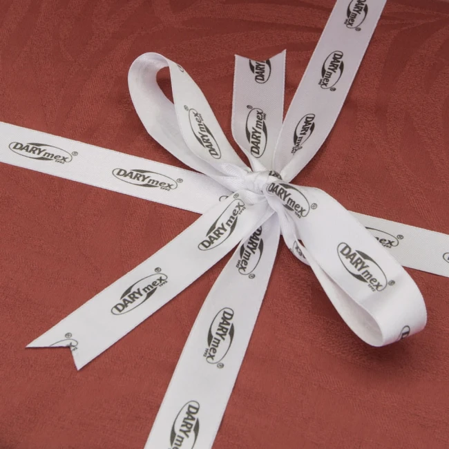 Zbliżenie na prezent zapakowany w ciemnoczerwony papier, przewiązany białą wstęgą z nadrukiem DARYmex. Wstążka utworzona w kokardę z dwoma długimi końcami, dodając elegancki akcent do opakowania. Tło proste, jednolite.