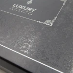 Opakowanie pościeli satynowej w kolorze ciemnoszarym z widocznym, wypukłym wzorem w stylu żakardowym. Na górze znajduje się napis LUXURY COLLECTION w eleganckim kroju, otoczony dekoracyjnym elementem. Opakowanie ma prostokątny kształt i jest zamknięte.