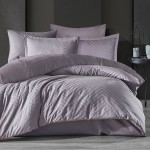 Pościel satyna bawełniana żakardowa 220x200 w odcieniach fioletu, leżąca w złożeniu. Poduszki w zestawie mają delikatny wzór w formie linii, a całość ułożona na szarym tle. W widocznym tle czarna metalowa dekoracja oraz elementy aranżacji.