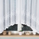 Firana żakardowa Bakaro w kolorze białym, o wymiarach 420x160 cm, z dekoracyjnym wzorem roślinnym na dolnej krawędzi, wisząca na oknie. Tło stanowi drewniana parapet z doniczką i świątecznym kalendarzem, co tworzy aranżację domową.