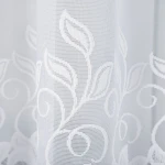 Biała firana żakardowa Bastia o wymiarach 500x160 cm, z delikatnym wzorem liści i spiral. Ujęcie zbliżeniowe, ukazujące fakturę i szczegóły wzoru. Podkreśla lekkość materiału, zawieszona, na jasnym tle.
