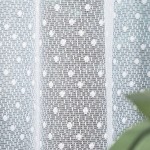 Firana gotowa Demetria żakardowa biała 400x160; ażurowa, biała tkanina z delikatnym wzorem kwiatowym; wisząca na białym tle, ukazująca teksturę materiału; widok frontalny. Idealna dekoracja okien, wprowadzająca lekkość do wnętrza.