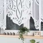 Biała firana żakardowa o wymiarach 500x160 cm, z motywem roślinnym, wisząca w oknie. Tło z zewnątrz przedstawia zieleń, a na parapecie znajdują się ozdobne elementy: doniczka z rośliną i dekoracyjne kostki z napisem HOME oraz datą.