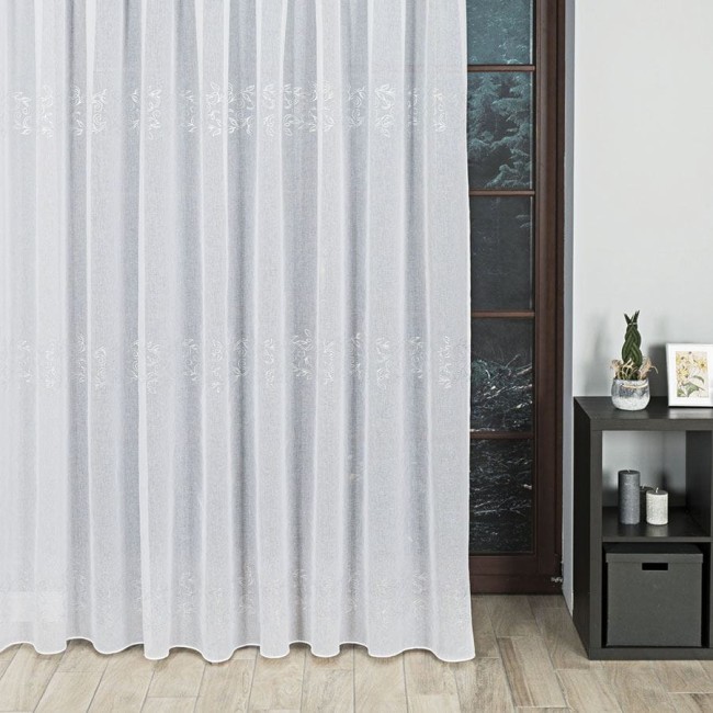 Firana fantazyjna biała z wzorem ołowianka, wysokość 290 cm, zawieszona w oknie. Materiał lekki i zwiewny, z delikatnym, haftowanym wzorem. Widoczna elegancka faktura. Tło w odcieniu szarości, meble w minimalistycznej aranżacji.