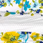 Pościel w niebieskie i żółte kwiaty 160x200 Tual z mikrowłókna, rozłożona na neutralnym tle. Wzór kwiatowy dominuje na białym tle, z widocznymi detalami w postaci dużych, kolorowych kwiatów oraz drobnych kropek.