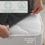 Prześcieradło jersey z elastanem w kolorze czarnym, rozłożone na materacu o grubości 25 cm, z widoczną elastyczną gumką. Zdjęcie wykonane z boku, pokazujące detal zakładania prześcieradła. Na tle jasnego materaca i otoczenia w neutralnych barwach.