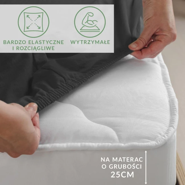 Prześcieradło jersey z elastanem w kolorze czarnym, rozłożone na materacu o grubości 25 cm, z widoczną elastyczną gumką. Zdjęcie wykonane z boku, pokazujące detal zakładania prześcieradła. Na tle jasnego materaca i otoczenia w neutralnych barwach.