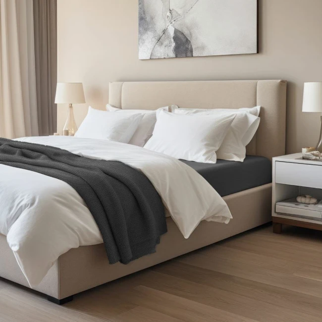 Prześcieradło jersey z elastanem w kolorze czarnym, o wymiarach 160x200 cm, rozłożone na łóżku. Widoczna struktura materiału, leżące białe poduszki i koc w ciemnym kolorze. Otoczenie z jasną ścianą i nowoczesnym nocnym stolikiem.