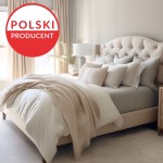 Prześcieradło jersey z elastanem z gumką o wymiarach 200x220 cm, w kolorze białym, leżące na łóżku. Tło jasne, z dekoracyjnymi poduszkami w odcieniach beżu i szarości. Widoczne zasłony w jasnych tonacjach oraz lampa nocna obok łóżka.