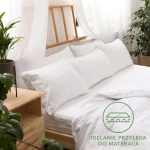 Prześcieradło jersey z gumką 90x200 w kolorze białym, rozłożone na łóżku. Kąt ujęcia z boku, ukazujący gładką strukturę materiału i gumkę przylegającą do materaca. Tło z naturalnymi roślinami i drewnianym meblem, co tworzy przytulny kontekst.