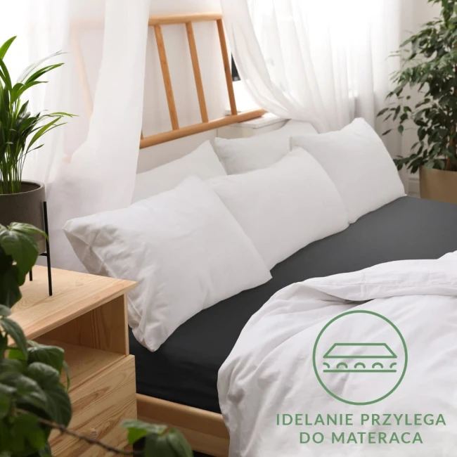 Prześcieradło jersey z gumką 160x200 w kolorze czarnym, rozłożone na łóżku, idealnie przylegające do materaca. Widoczne białe poduszki w tle, drewniane elementy mebli oraz roślina doniczkowa. Tło oświetlone naturalnym światłem z zasłonami.