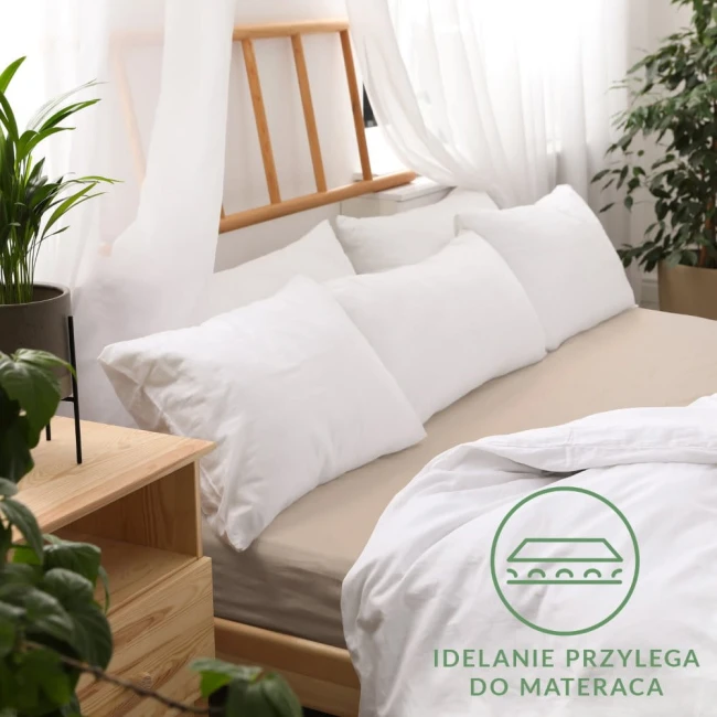 Prześcieradło jersey z gumką w kolorze beżowym, rozmiar 120x200 cm, leżące na łóżku z widoczną narzutą i białymi poduszkami. Tło w jasnych, naturalnych kolorach, z dodatkowymi roślinami doniczkowymi. Widoczne logo w kolorze zielonym w dolnym rogu.