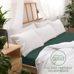 Prześcieradło jersey z gumką 180x200 w kolorze butelkowej zieleni, leżące na łóżku. Widoczne białe poduszki oraz jasnożółta deska łóżka. Tło naturalne z roślinnością i zasłonami, podkreślające elegancką aranżację. Idealnie przylega do materaca.