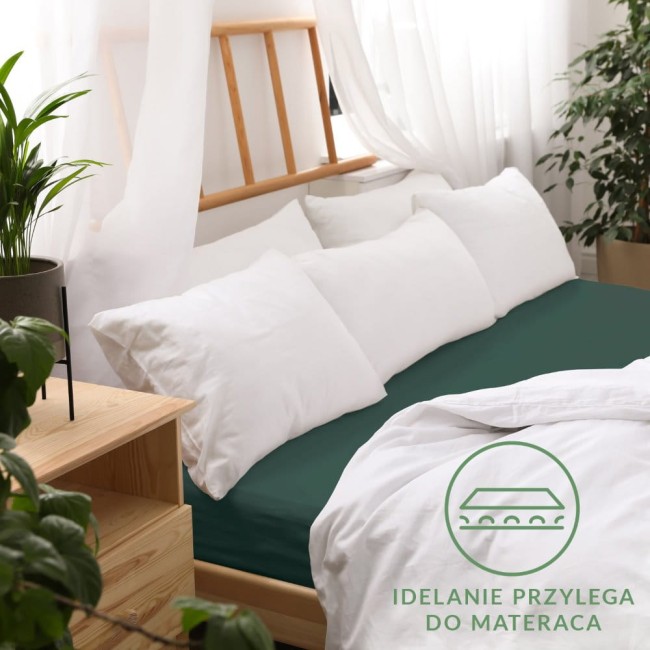 Prześcieradło jersey z gumką 180x200 w kolorze butelkowej zieleni, leżące na łóżku. Widoczne białe poduszki oraz jasnożółta deska łóżka. Tło naturalne z roślinnością i zasłonami, podkreślające elegancką aranżację. Idealnie przylega do materaca.