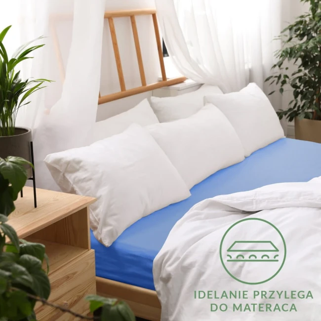 Prześcieradło jersey z gumką 160x200 w kolorze chabrowym, leżące na łóżku, idealnie przylega do materaca. Otoczenie to jasne poduszki oraz drewniane elementy mebli. Widoczne napisy na materiale z informacją o dopasowaniu.