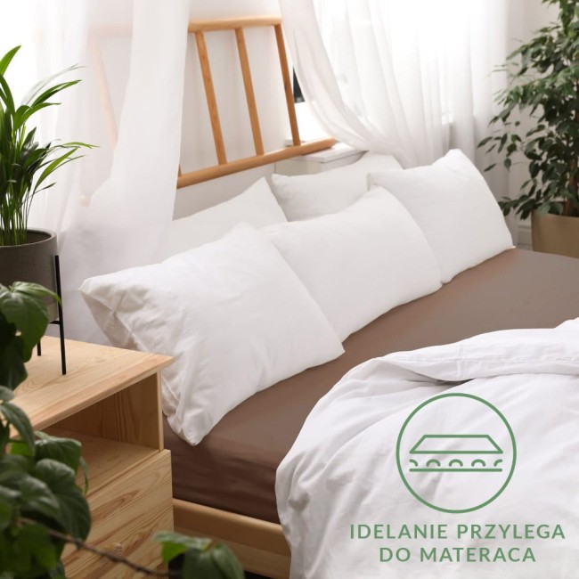 Prześcieradło jersey z gumką 90x200 w kolorze czekoladowym na łóżku, z białymi poduszkami ułożonymi wzdłuż. Produkt leży na rozłożonym materacu, z delikatnym białym prześcieradłem częściowo odsłoniętym. W tle widać rośliny oraz drewniane elementy mebli.