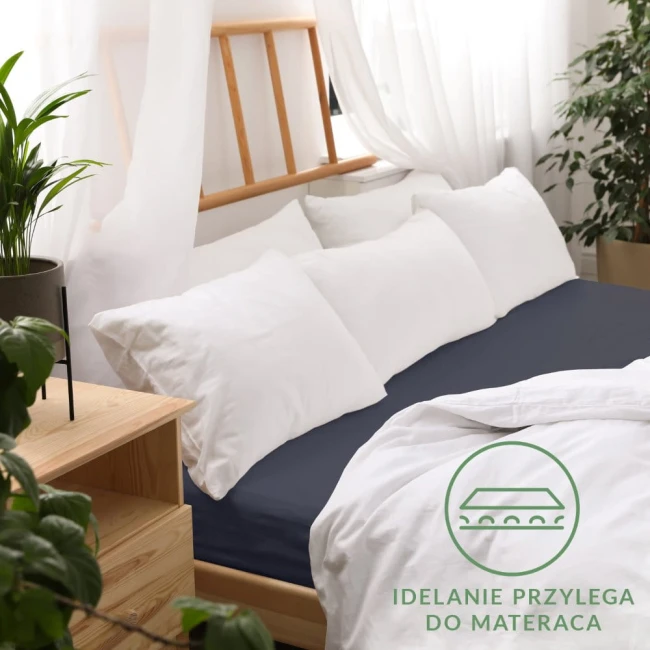 Prześcieradło jersey z gumką w ciemnoniebieskim kolorze, rozmiar 120x200 cm, umieszczone na łóżku z białą pościelą i poduszkami. Prześcieradło idealnie przylega do materaca, widoczne logo z przesłaniem o dopasowaniu. Tło jasne i naturalne.