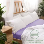 Jasnofioletowe prześcieradło jersey z gumką, rozmiar 200x220 cm, leży na materacu, otoczone białymi poduszkami i pościelą. Widoczne naturalne tło z drewnianym łóżkiem oraz roślinami. Z przodu widoczny napis idealnie przylega do materaca.