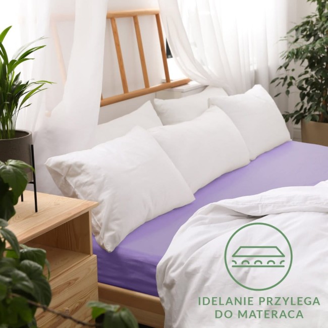 Jasnofioletowe prześcieradło jersey z gumką, rozmiar 200x220 cm, leży na materacu, otoczone białymi poduszkami i pościelą. Widoczne naturalne tło z drewnianym łóżkiem oraz roślinami. Z przodu widoczny napis idealnie przylega do materaca.