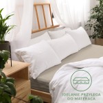 Prześcieradło jersey z gumką 180x200 w kolorze Kakao leży na łóżku z jasnym, drewnianym wezgłowiem, otoczone białymi poduszkami. Widoczna jest struktura materiału.