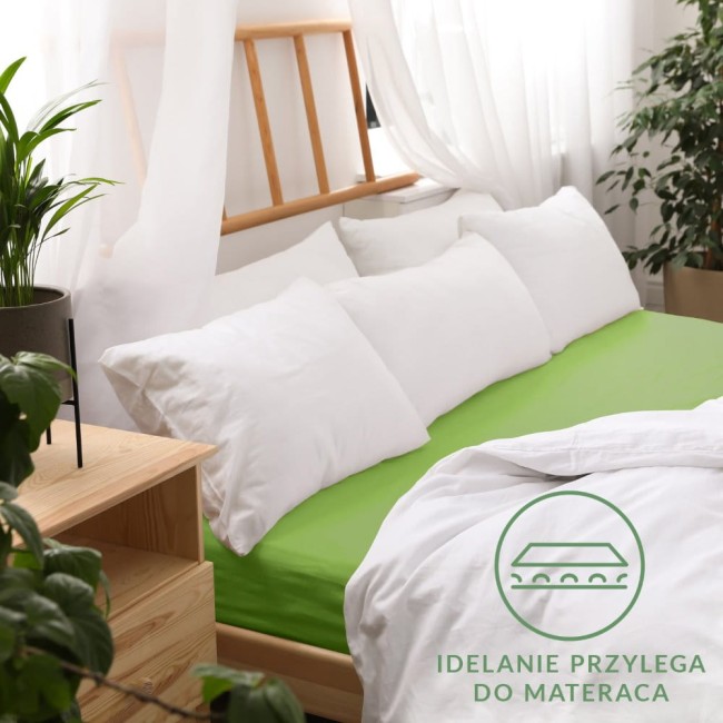 Prześcieradło jersey z gumką w kolorze kaktusowym, w rozmiarze 140x200 cm, leży na łóżku otoczonym białymi poduszkami. Widoczna struktura materiału oraz gumka stabilizująca. W tle jasne firany i rośliny doniczkowe, co tworzy przytulny klimat.