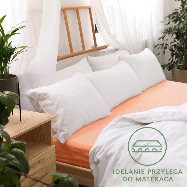 Prześcieradło jersey z gumką w kolorze łososiowym, o wymiarach 200x220 cm, leży na łóżku, podparte na białych poduszkach.