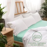 Prześcieradło jersey z gumką w kolorze morskim, rozmiar 90x200, leży na łóżku z białymi poduszkami. Widoczna faktura materiału, idealnie przylega do materaca. W tle roślinność i naturalne światło tworzą przytulną atmosferę.