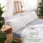 Prześcieradło jersey z gumką w kolorze popielatym, rozmiar 180x200 cm, rozłożone na materacu. W tle widoczne poduszki w białych poszewkach oraz drewniane łóżko, otoczone roślinnością. Na zdjęciu widoczny zestaw białych poduszek.