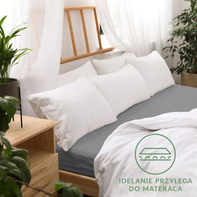 Prześcieradło jersey z gumką 120x200 w kolorze szarym leży na łóżku, obok białych poduszek. Widoczny jest detal gumki, która zapewnia idealne przyleganie do materaca. Tło jest jasne z naturalnymi akcentami roślinnymi.