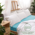 Prześcieradło jersey z gumką 80x160 w kolorze turkusowym, rozłożone na łóżku z białą pościelą. Kąt ujęcia frontalny, z wyraźnym widokiem na elastyczność materiału. Tło jasne z dekoracyjną roślinnością i drewnianymi meblami.