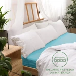 Prześcieradło jersey z gumką 120x200 w turkusowym kolorze, rozłożone na łóżku z białymi poduszkami w tle. Widoczna faktura materiału oraz elastyczna gumka. Tło jasne, z elementami drewnianymi i roślinnymi. Produkt idealnie przylega do materaca.