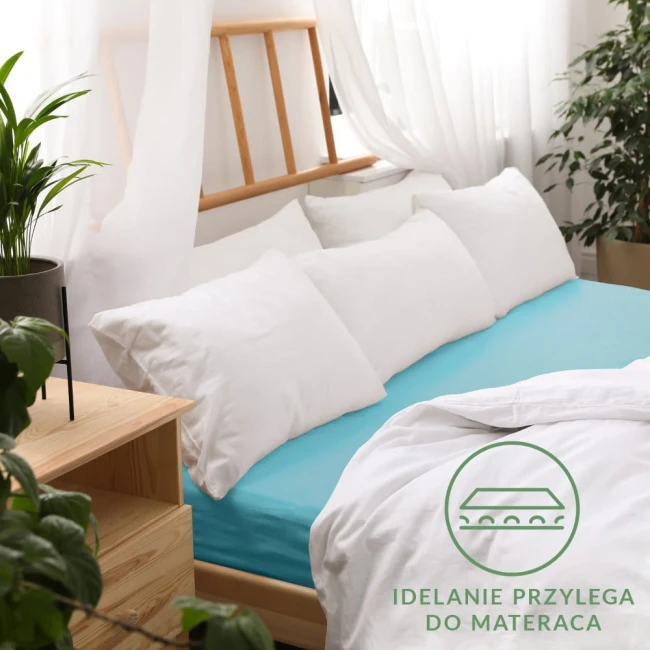 Prześcieradło jersey z gumką 120x200 w turkusowym kolorze, rozłożone na łóżku z białymi poduszkami w tle. Widoczna faktura materiału oraz elastyczna gumka. Tło jasne, z elementami drewnianymi i roślinnymi. Produkt idealnie przylega do materaca.
