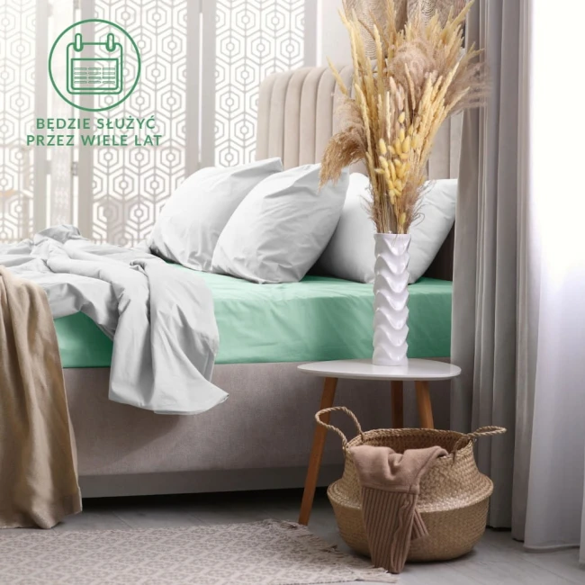 Prześcieradło frotte z gumką 160x200 w kolorze morskim, rozłożone na łóżku z poduszkami, leżące na jasnoszarym materacu. W tle widoczna elegancka dekoracja okna oraz minimalistyczny stolik z białym wazonem i suszonymi kwiatami.