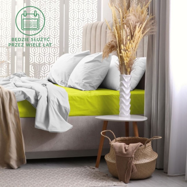 Prześcieradło frotte z gumką w kolorze limonka o wymiarach 200x220 cm leży na łóżku, otoczone białymi poduszkami. Tło stanowi nowoczesna aranżacja sypialni z drewnianą podłogą i jasnymi dekoracjami.