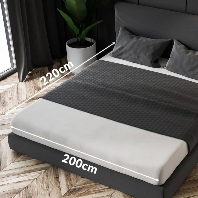 Prześcieradło satynowe z gumką w kolorze czarnym, w rozmiarze 200x220 cm, leży na łóżku. Ujęcie z góry ukazuje fakturę materiału i podkreśla nici w kontrastującym kolorze. W tle widoczne poduszki oraz roślina w doniczce, na tle jasnej podłogi.