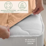 Prześcieradło jersey z elastanem w kolorze różowym, rozłożone na materacu. Kąt ujęcia zbliżony z ręką chwytającą krawędź. Widoczne napisy informujące o elastyczności i trwałości oraz o dopasowaniu do materaca o grubości 25 cm. Tło rozmyte, neutralne.