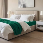 Prześcieradło jersey z elastanem w kolorze zielonym o wymiarach 140x200, rozłożone na łóżku z białą pościelą. Produkt widoczny z boku, z detalami o teksturze materiału i gumce mocującej. Otoczenie nowoczesnej sypialni z jasnymi kolorami.