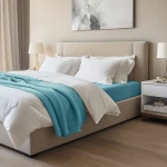Prześcieradło jersey z elastanem 180x200 w kolorze niebieskim, widoczne na materacu łóżka, z lekko podniesioną krawędzią. Otoczenie to jasna sypialnia z beżową ramą łóżka i białą pościelą. Na wierzchu leży turkusowy koc.