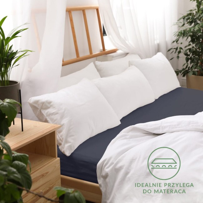 Prześcieradło jersey z gumką 160x200 w kolorze ciemnego granatu, rozłożone na łóżku z białymi poduszkami i pościelą. Ujęcie z boku, pokazujące gładką fakturę materiału.