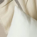 Poszewka dekoracyjna 45x45 w kolorze białym, gładka, z widoczną teksturą materiału. Ujęcie z góry, ukazujące narożnik poduszki. Produkt leży na jasnym tle obok tkaniny, co podkreśla jego kolor i fakturę.